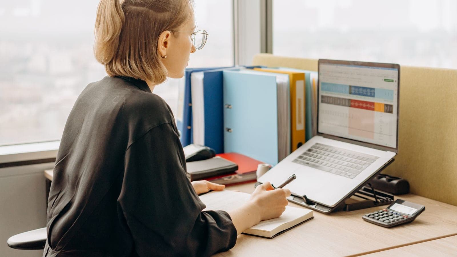 Account manager at a desk with a client spreadsheet open on a laptop and a shelf of client binders behind